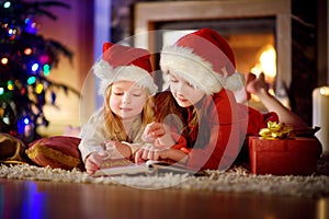 Two cute little sisters reading a story book together under a Christmas tree