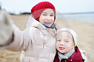 Two cute little sisters taking a picture of themselves at winter beach on cold winter day. Kids playing by the ocean.