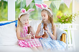 Two cute little sisters wearing bunny ears eating chocolate Easter rabbits. Kids playing egg hunt on Easter. Children celebrate Ea