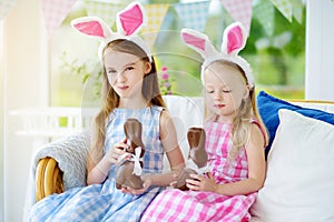 Two cute little sisters wearing bunny ears eating chocolate Easter rabbits. Kids playing egg hunt on Easter.