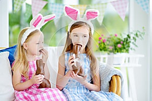 Two cute little sisters wearing bunny ears eating chocolate Easter rabbits. Kids playing egg hunt on Easter.