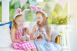 Two cute little sisters wearing bunny ears eating chocolate Easter rabbits. Kids playing egg hunt on Easter.