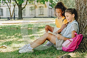 Cute multicultural schoolgirls sitting on lawn under tree and reading book together
