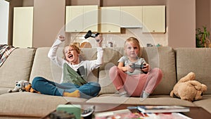 Two cute siblings, little boy and girl playing video games, sitting on a couch in the living room