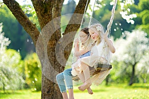 Two cute sisters having fun on a swing in blossoming old apple tree garden outdoors on sunny spring day.