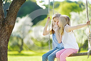 Two cute sisters having fun on a swing in blossoming old apple tree garden outdoors on sunny spring day