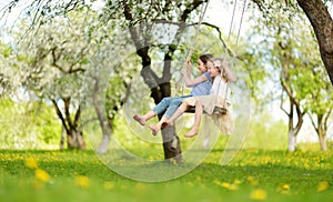 Two cute sisters having fun on a swing in blossoming old apple tree garden outdoors on sunny spring day