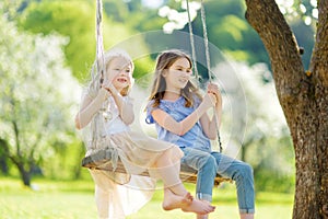 Two cute sisters having fun on a swing in blossoming old apple tree garden outdoors on sunny spring day