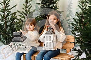 Two cute sisters are sitting in a Christmas interior and holding lanterns.