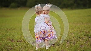 Two cute sisters twins frolicking on a green meadow in summer day. Holding hands smile at each other.