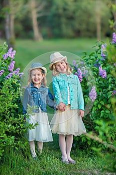 Two cute smiling girls sisters lovely together on a lilac field bush all wearing stylish dresses and jeans coats.