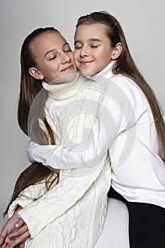 Two cute teenage girlfriends schoolgirls wearing white turtleneck sweaters, smiling sit, hugging each other in a friendly way.