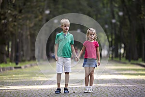 Two cute young blond smiling children, girl and boy, brother and sister holding hands on blurred bright sunny summer park alley