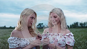 Two cute young blonde women with tattooes standing on a field holding daisies