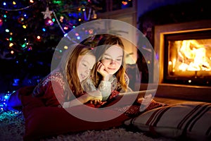 Two cute young sisters using a tablet pc at home by a fireplace in warm and cozy living room on Christmas eve