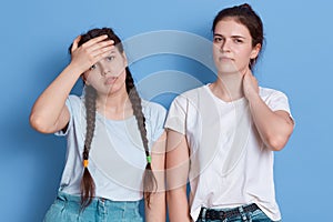 Two dark haired sisters posing indoors against blue wall, sad girl with health problems, stressed female colleague has pain in