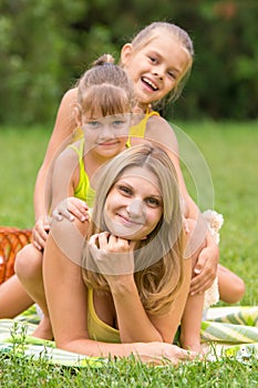 Two daughters are sitting on young mother, who is lying on the green grass