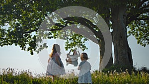 Two daughters walking along green field to their mother, swinging on a rope swing tied to old tree.