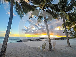 Two deck chairs under palm trees at sunset on an empty beach at