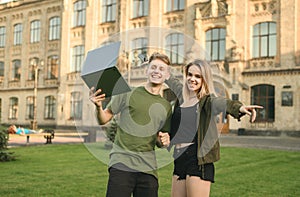 Two delighted happy students laughing at the college park standing with the laptop on the grass. Cheerful students couple in front