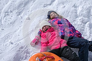 Two delightful laughing kids girls wearing ski suit lying on white snow with rubber tube during winter season travel