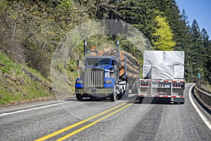 Two different big rigs semi trucks with loaded semi trailers move in opposite directions on a narrow winding road in the forest