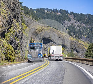 Two different classic loaded big rigs semi trucks with semi trailers moving in opposite directions on the narrow winding road with