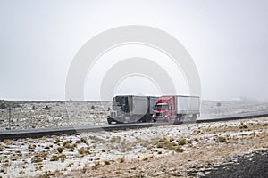 Two different long hauler big rigs semi trucks with dry van semi trailers running side by side on the winter highway road at