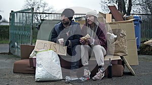 Two dirty and poorly dressed homeless people, a man and a woman, sit by a pile of rubbish with a handwritten CRISIS