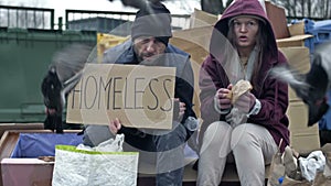 Two dirty and poorly dressed homeless people, a man and a woman, sit by a pile of rubbish with a handwritten HOMELESS