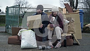 Two dirty and poorly dressed homeless people, a man and a woman, sit by a pile of rubbish with a handwritten HUNGRY