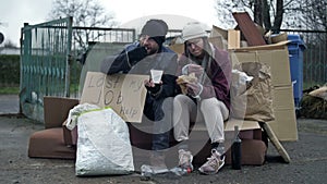 Two dirty and poorly dressed homeless people, a man and a woman, sit by a pile of rubbish with a handwritten LAST MY JOB