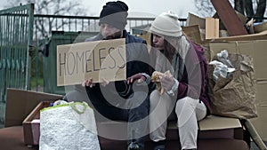Two dirty and poorly dressed homeless people, a man and a woman, are sitting near a pile of rubbish with a handwritten