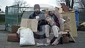 Two dirty and poorly dressed homeless people, a man and a woman, are sitting near a pile of rubbish with a handwritten