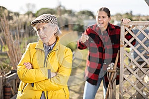 Two disappointed young and old female neighbors quarreling while standing near fence on garden during daytime in March