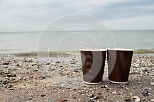 Two disposable paper cups of coffee standing on sand against blue sea, copy space. Take-out coffee