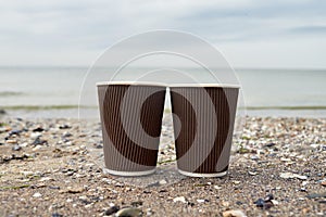 Two disposable paper cups of coffee standing on sand against blue sea, copy space. Take-out coffee