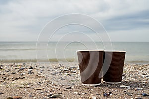 Two disposable paper cups of coffee standing on sand against blue sea, copy space. Take-out coffee
