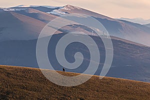 Two distant people on a mountain, with snowcapped peaks and wind turbines