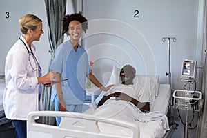 Two diverse female doctors and african american male patient in hospital room smiling to each other