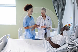 Two diverse female doctors and african american male patient in hospital room talking