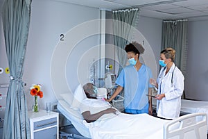 Two diverse female doctors wearing masks standing next to male patient in hospital room