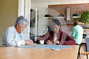 Two diverse female friends sitting in kitchen with coffee and doing puzzles