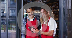 Two diverse male baristas wearing aprons standing in the doorway and using digital tablet