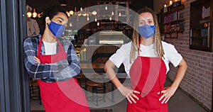 Two diverse male baristas wearing face masks and aprons standing in doorway of cafe
