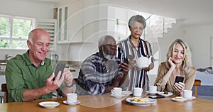 Two diverse senior couples sitting by a table drinking tea using smartphones at home