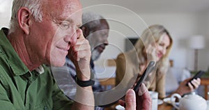 Two diverse senior couples sitting by a table drinking tea using smartphones at home