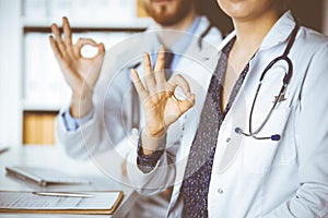 Two doctors sitting with Ok sign in hospital office. Medical help, countering viral infection and medicine concept