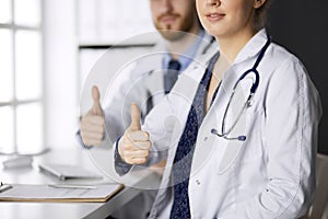 Two doctors sitting with thumbs up in hospital office. Medical help, countering viral infection and medicine concept