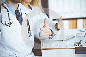 Two doctors sitting with thumbs up in hospital office. Medical help, countering viral infection and medicine concept